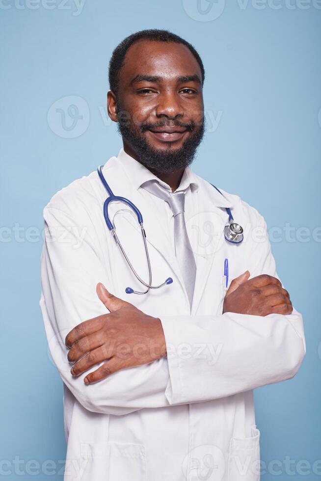 sorridente medico di africano americano etnia avendo il suo braccia attraversato è posizionato nel davanti di isolato blu sfondo. maschio assistenza sanitaria specialista con stetoscopio in posa per telecamera nel studio. foto