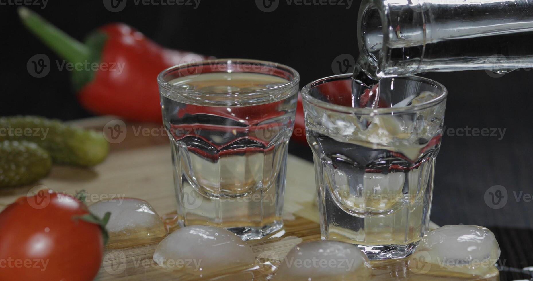 versare Vodka a partire dal un' bottiglia in tiro bicchieri con ghiaccio cubi su il tavolo foto