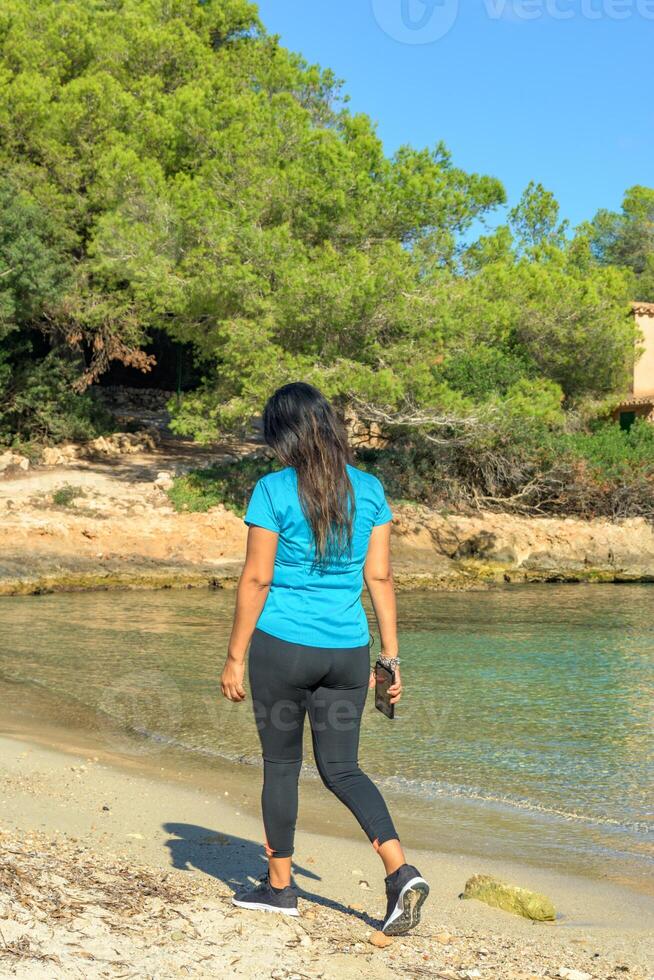 latina donna nel abbigliamento sportivo a piedi e ascoltando per musica con sua inteligente Telefono su il spiaggia foto