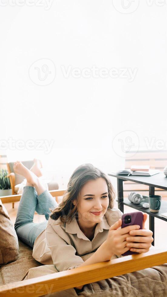 allegro del Millennio donna rilassante su divano, guardando a Telefono, lettura messaggi, chattare, e godendo gratuito volta. accogliente tempo libero a casa con contento giovane donna coinvolgente nel digitale comunicazione. foto