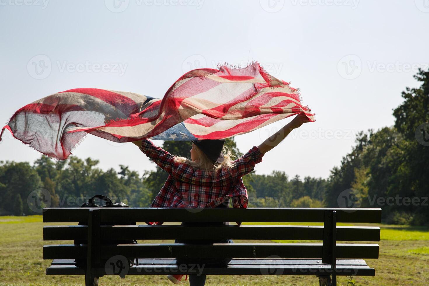 giovane ragazza si siede su un' panchina e detiene un' scialle con un' modello di il americano bandiera nel sua mani agitando nel il vento. alto qualità foto