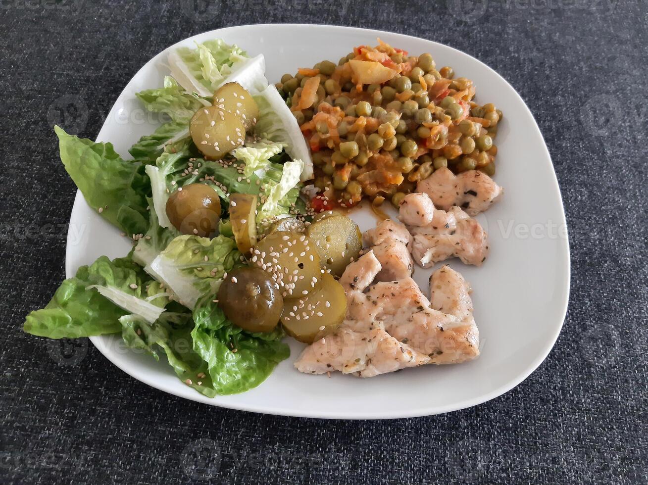 fatti in casa grigliato pollo con verde insalata, piselli stufato e cetriolo sottaceti servito su un' bianca piatto foto