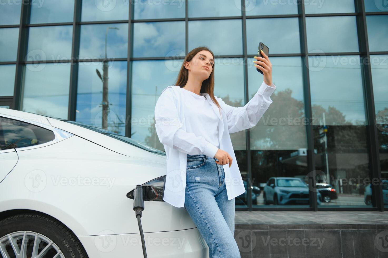 Telefono nel mani. donna su il elettrico macchine caricare stazione a giorno. marca nuovo veicolo. foto