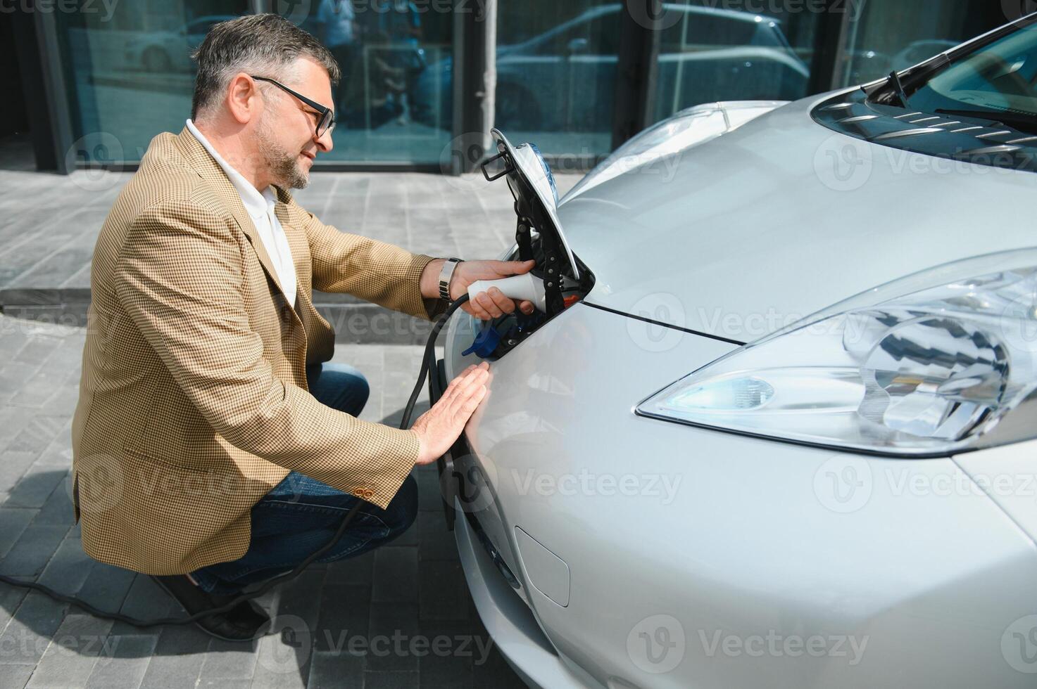 bello barbuto uomo seduta vicino il suo nuovo moderno elettrico auto e Tenere spina di il caricabatterie, mentre auto è ricarica a il ricarica stazione foto