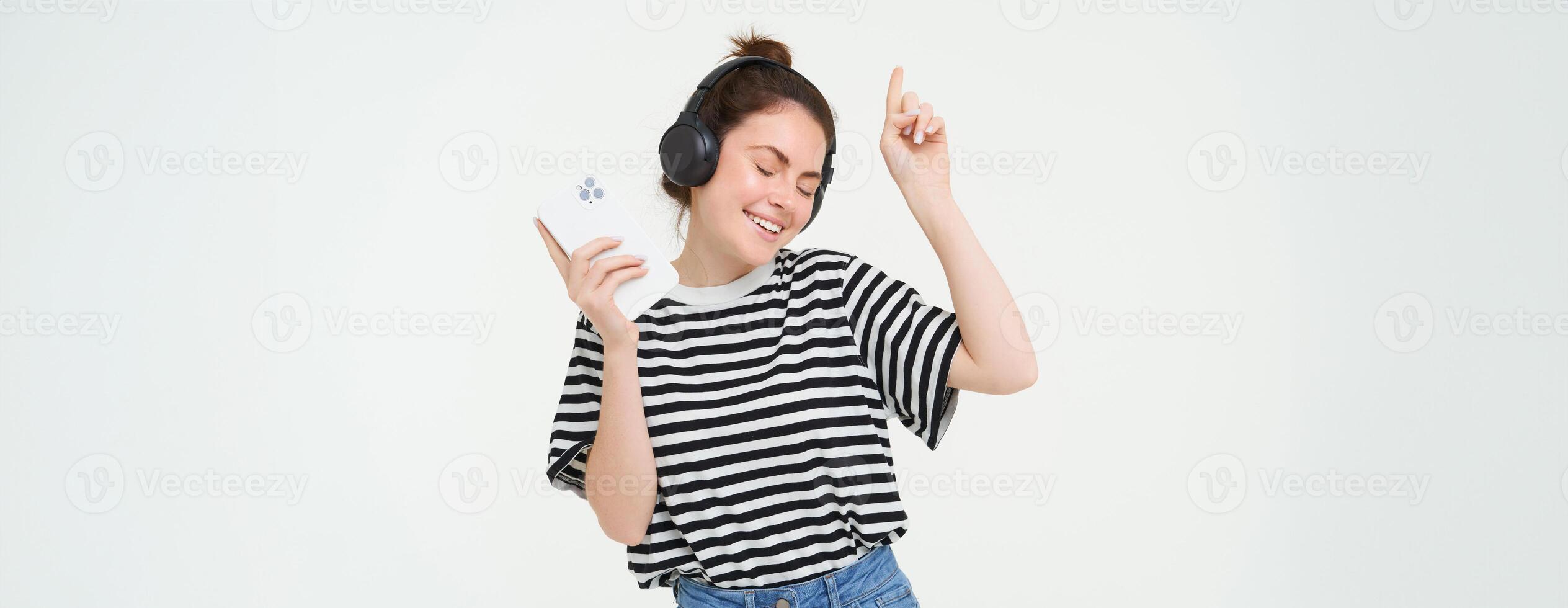 ritratto di contento donna con smartphone i cambiamenti canzone su mobile Telefono streaming app, ascolta musica nel cuffia, danza contro bianca sfondo foto