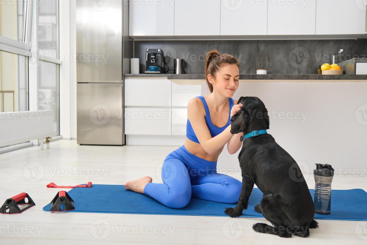 Immagine di giovane donna fa allenarsi a partire dal casa, si siede su yoga stuoia nel vivente camera e giochi con sua nero cane, cucciolo distrae ragazza a partire dal fare esercizi foto