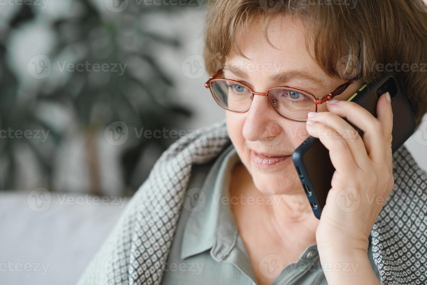 bellissimo anziano donna nel casuale capi di abbigliamento utilizzando sua inteligente Telefono mentre seduta su il divano a casa foto