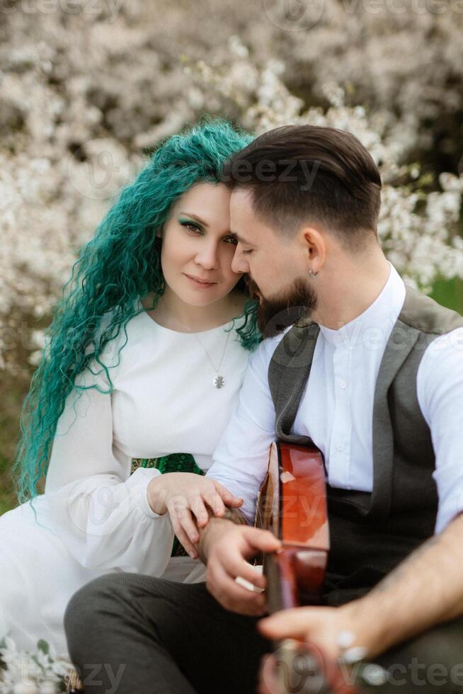 un' barbuto sposo giochi un' a corda strumento e un' ragazza con si siede nel un' primavera prato foto
