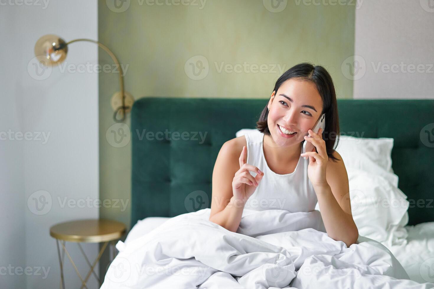 bellissimo giovane coreano donna chiamando, avendo un' Telefono chiamata nel letto, parlando con amico mentre soggiorno nel Camera da letto foto