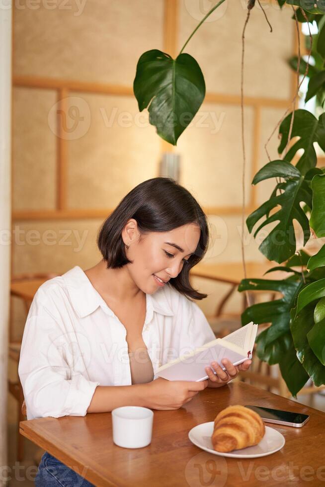 sognante giovane sorridente asiatico donna lettura prenotare, seduta nel bar, mangiare brioche e potabile caffè nel accogliente interno foto