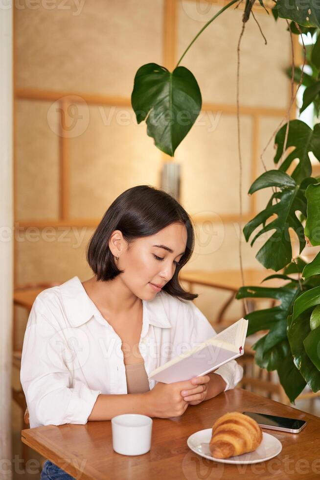 sognante giovane sorridente asiatico donna lettura prenotare, seduta nel bar, mangiare brioche e potabile caffè nel accogliente interno foto