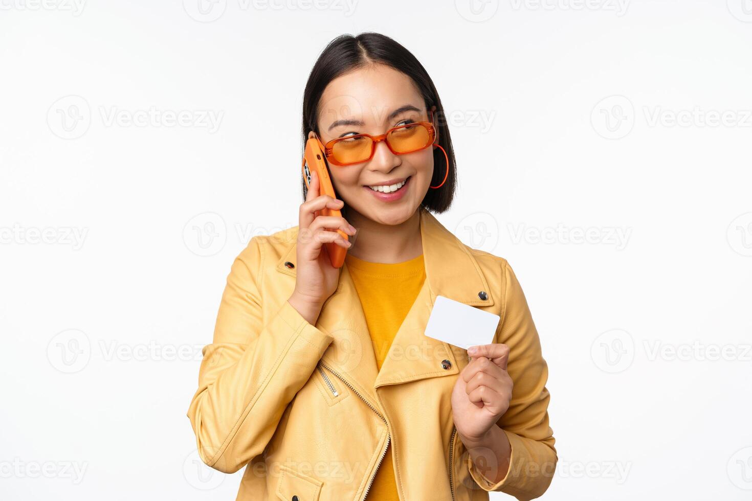 elegante asiatico femmina modello, parlando su smartphone e mostrando credito carta, in piedi al di sopra di bianca sfondo foto