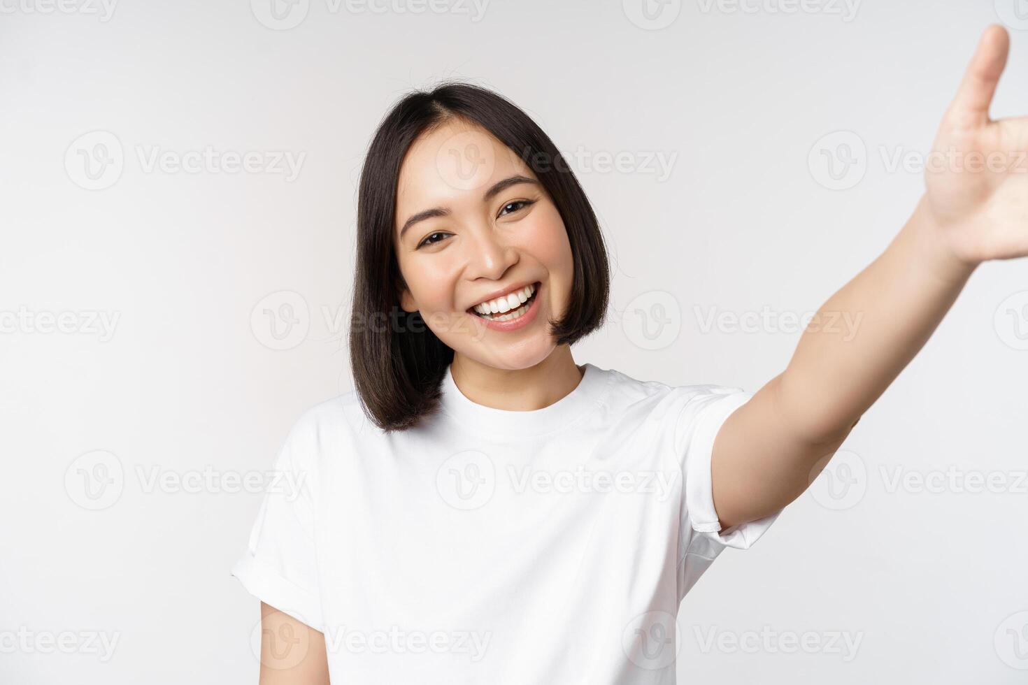 bellissimo giovane asiatico donna sorridente, guardare a telecamera, Tenere dispositivo, assunzione autoscatto, video Chiacchierare, in piedi nel maglietta al di sopra di bianca sfondo foto