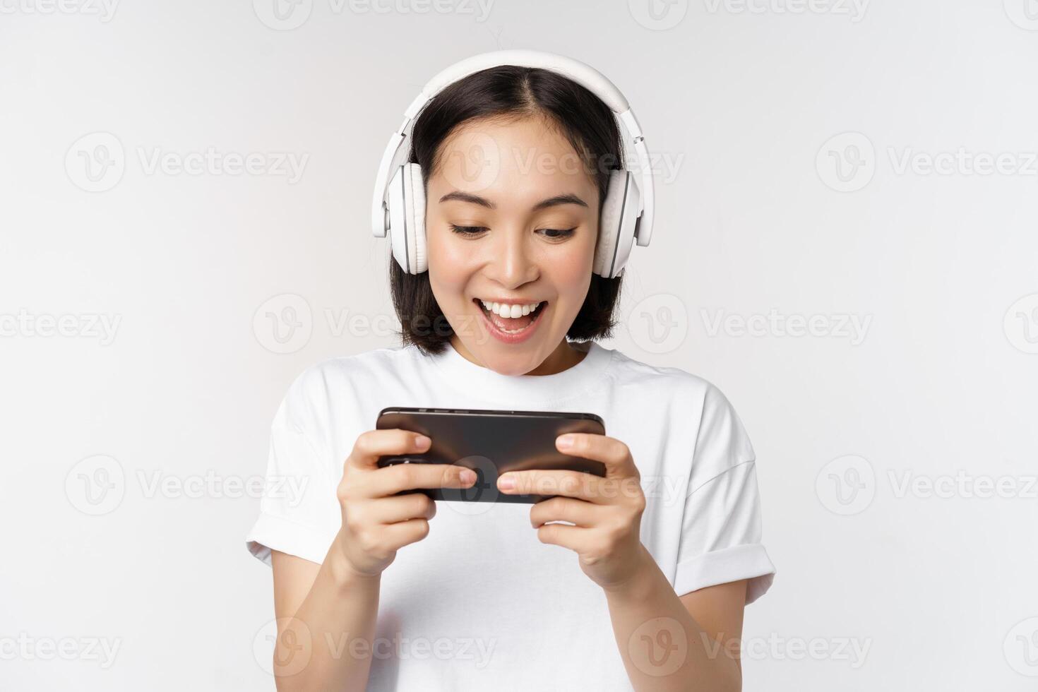 contento asiatico donna nel cuffia, guardare a smartphone, Guardando video su mobile Telefono e sorridente, in piedi al di sopra di bianca sfondo foto