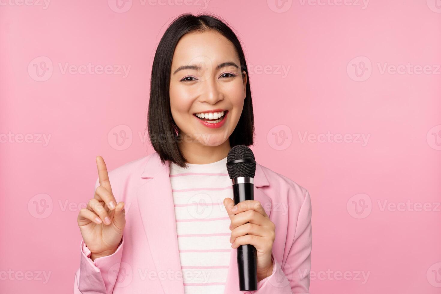 Immagine di entusiasta asiatico donna d'affari dando discorso, parlando con microfono, Tenere microfono, in piedi nel completo da uomo contro rosa studio sfondo foto