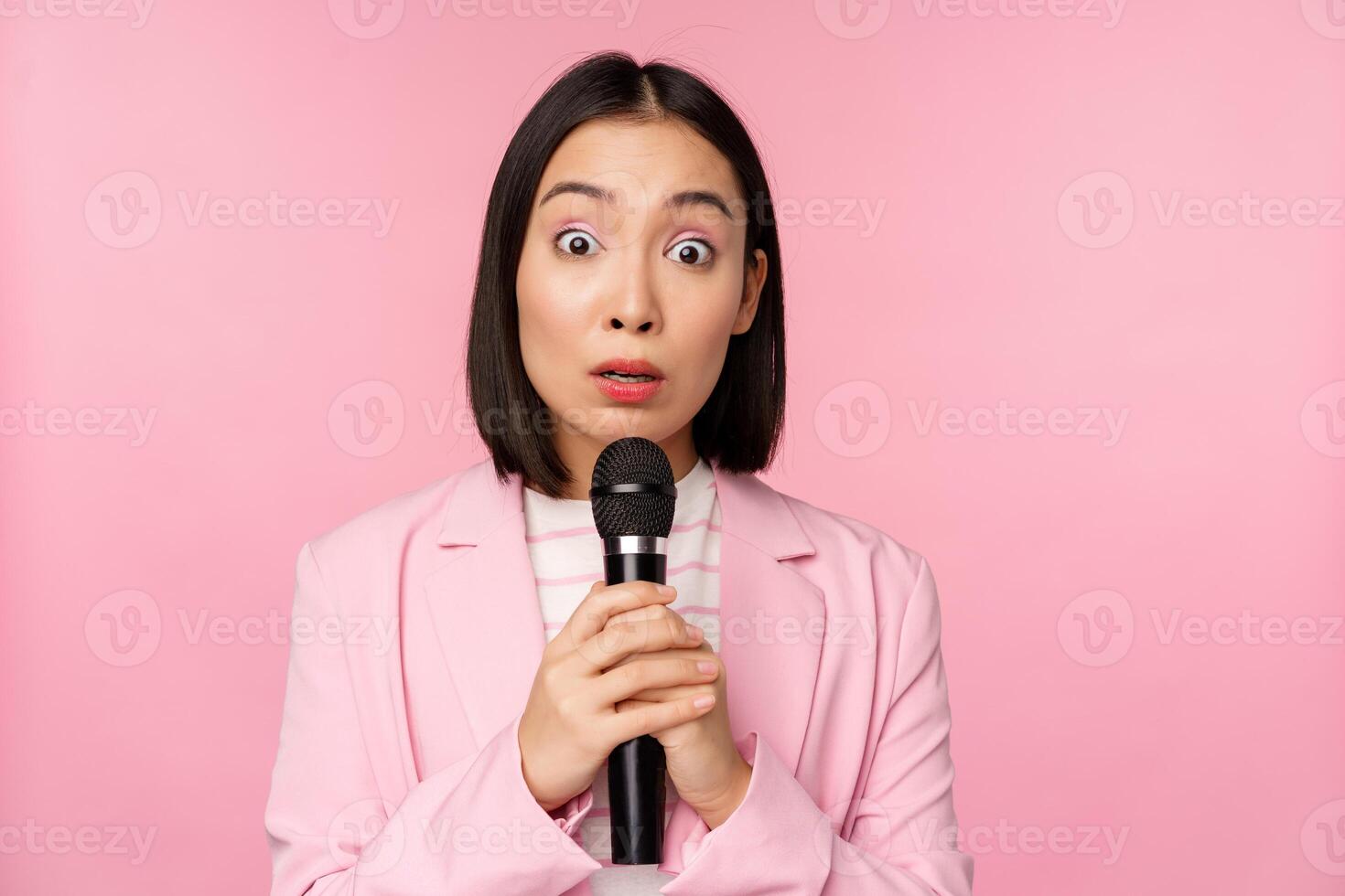 giovane asiatico commessa, ufficio signora con completo da uomo, Tenere microfono e guardare scioccato a telecamera, parlando, dando discorso, in piedi al di sopra di rosa sfondo foto