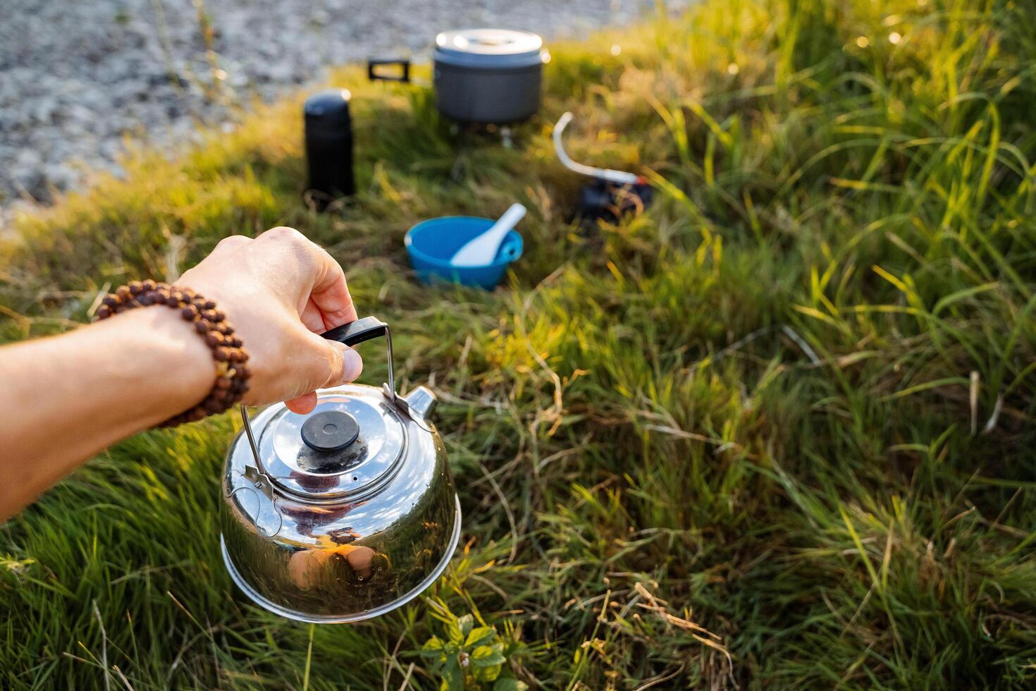 al affresco cenare. un ferro bollitore nel il mani di un' uomo in piedi vicino il verde banca di il fiume. messa a fuoco su il bollitore e mano, nel il sfondo il riposo di il cucina utensili. attivo riposo foto