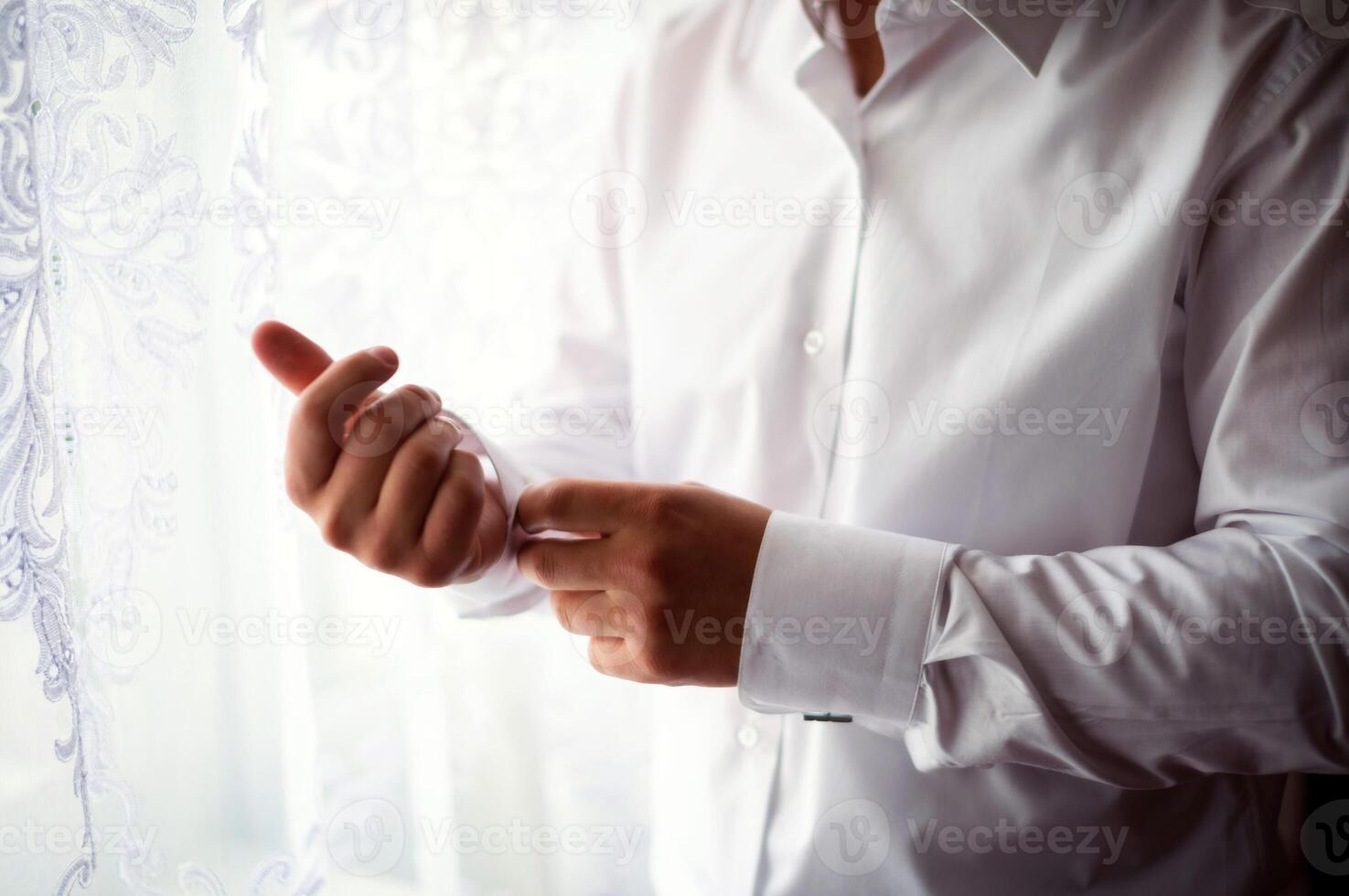 bello uomo pulsanti il suo bianca camicia in piedi a il finestra nel il mattina per nozze cerimonia. elegante uomo d'affari mette su manica camicia. avvicinamento foto