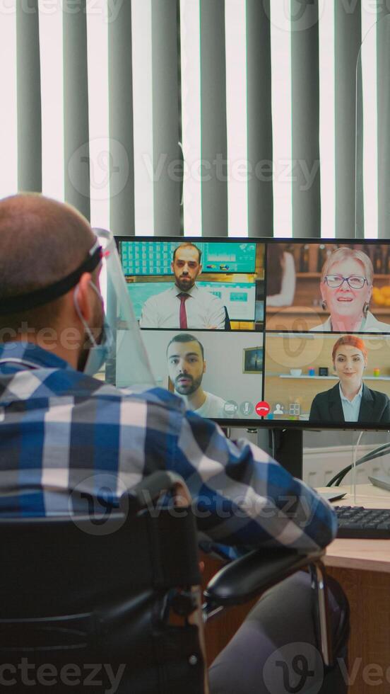 paralizzato manager nel sedia a rotelle parlando con maschera e visiera durante video chiamata avendo in linea conferenza nel attività commerciale ufficio. immobilizzato libero professionista Lavorando nel finanziario azienda rispetto sociale distanza foto