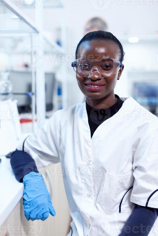 africano scienziato indossare protettivo bicchieri nel medico laboratorio guardare a telecamera. multietnico squadra di ricercatori Lavorando nel microbiologia laboratorio analisi soluzione per medico scopo. foto