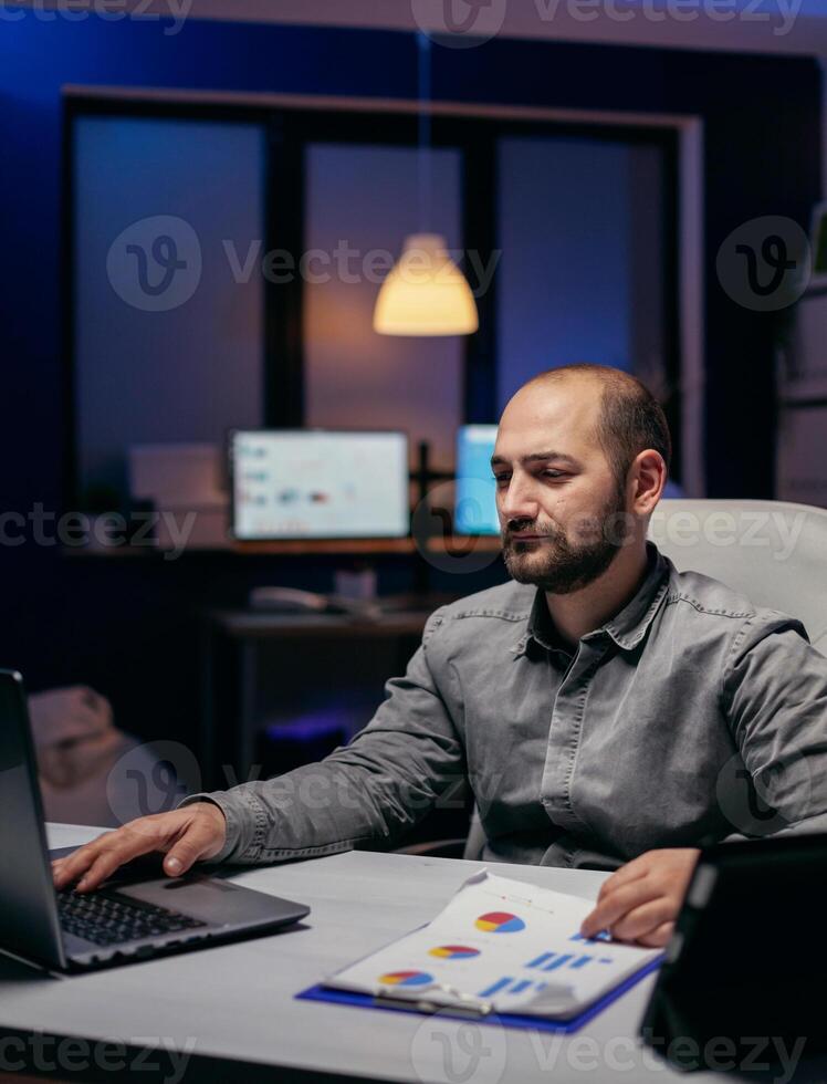 libero professionista Lavorando col tempo nel aziendale posto di lavoro guardare a il computer portatile seduta a scrivania. barbuto imprenditore fare sovraccarico a il ufficio nel il sera per finire un' Scadenza. foto