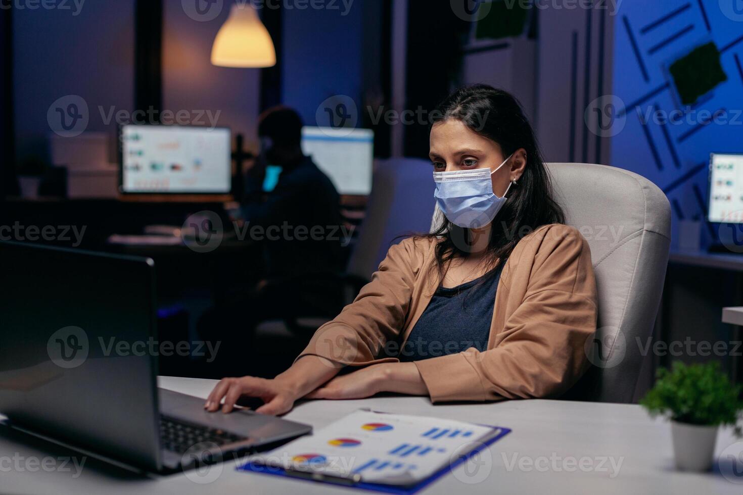 stressato caucasico giovane imprenditore fare col tempo indossare viso maschera . donna a seguire sociale distanziamento regole dovuto per coroanvirus pandemia mentre Lavorando in ritardo ore a il ufficio. foto