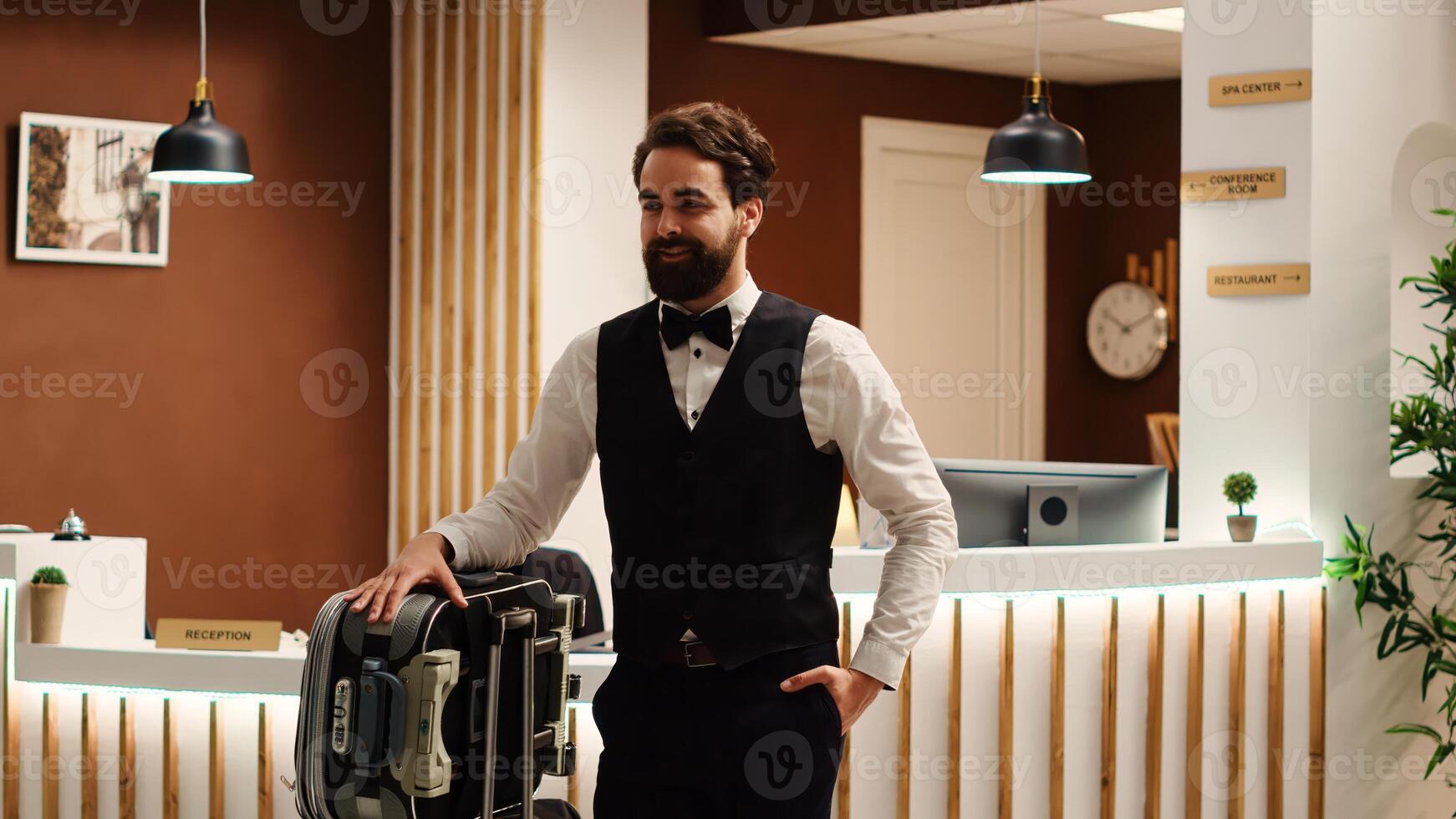 allegro sorridente professionale fattorino nel formale abbigliamento con Bagaglio carrello nel davanti di dai un'occhiata nel ricezione. contento Hotel dipendente trasporto bagaglio nel ricorrere atrio, svelare fiocco su tiro foto