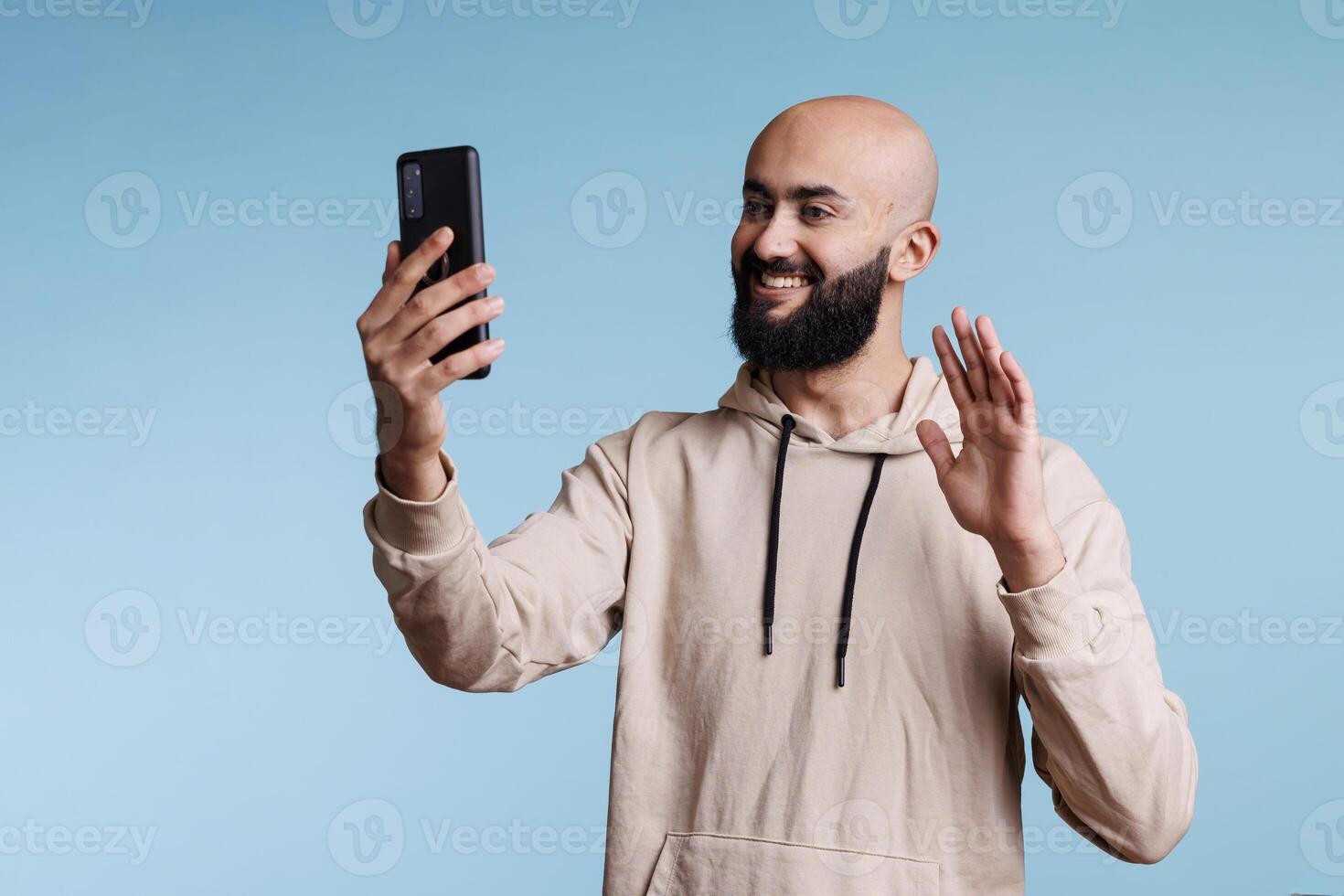 sorridente arabo uomo agitando Ciao mentre parlando in linea utilizzando smartphone video chiamata applicazione. amichevole persona avendo a distanza comunicazione, saluto con mano e A proposito di con mobile Telefono davanti telecamera foto