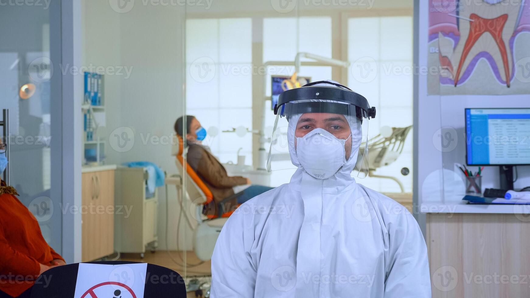 ritratto di stanco medico nel dentale ufficio guardare su telecamera indossare complessivamente e viso scudo seduta su sedia nel in attesa camera clinica. concetto di nuovo normale dentista visitare nel coronavirus scoppio. foto