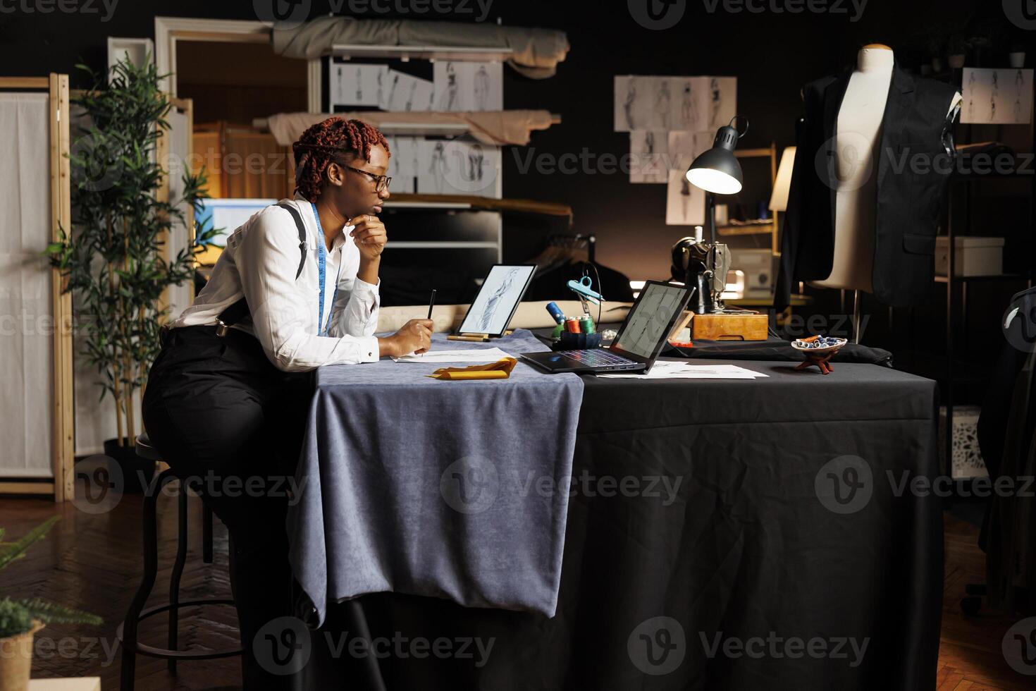 africano americano sarta nel Atelier negozio spazio di lavoro circondato di cucire strumenti guardare in linea per design ispirazioni, ispezionando schizzo disegni di imminente sartoriale collezione foto