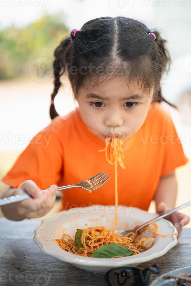 giovane ragazza bambini godendo un' forchettata di spaghetti. avvicinamento di un' poco ragazza nel un arancia camicia mangiare spaghetti con un' focalizzata espressione, all'aperto ristorante. foto