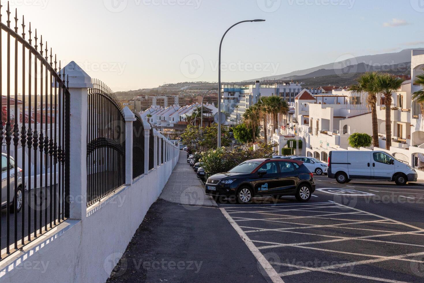 silenzioso Residenziale strada con parcheggiata macchine e bianca case, metallo recinto su il sinistra, e colline nel il sfondo a crepuscolo nel los cristiano, tenerife. foto