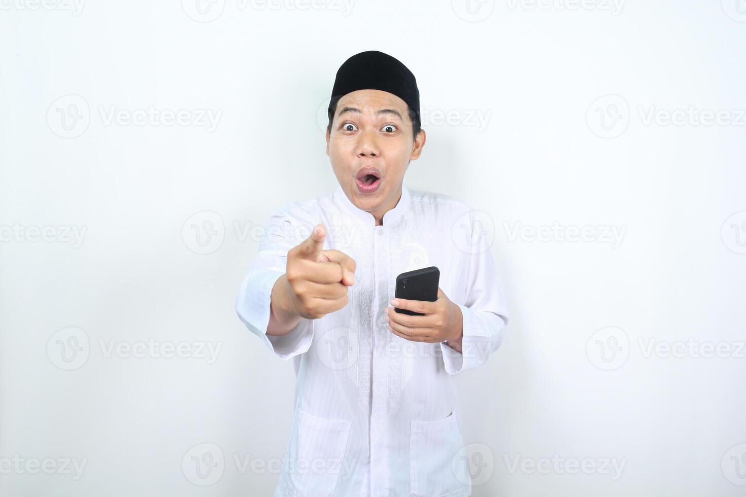 stupito asiatico musulmano uomo puntamento dito per telecamera con Tenere smartphone isolato foto