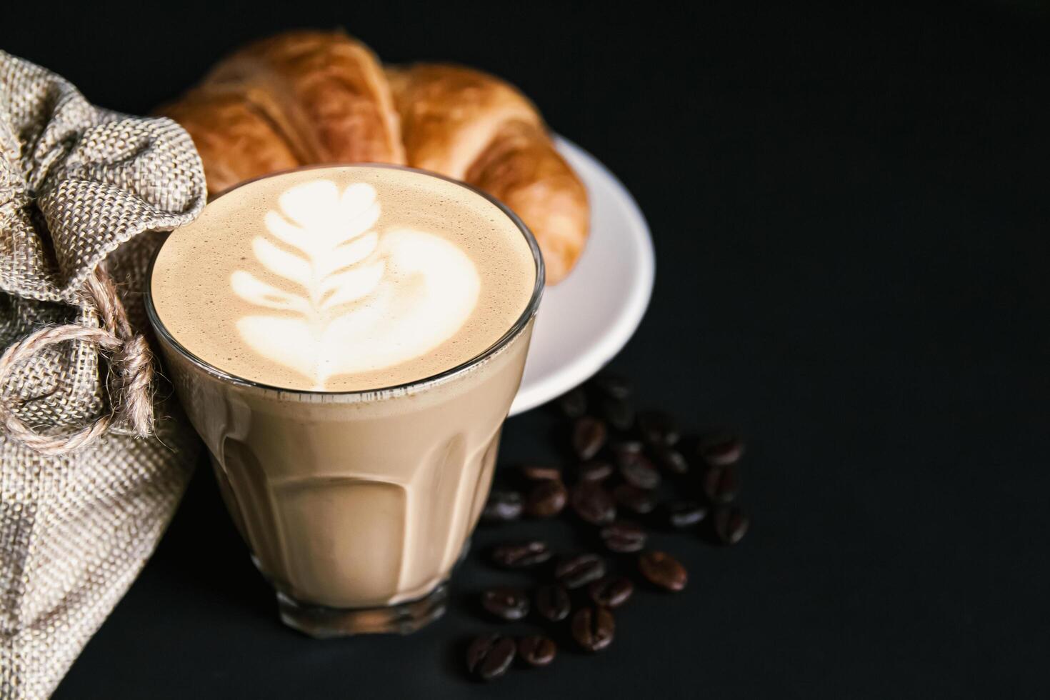 latte macchiato arte caffè nel un' bianca tazza, Cornetti, arrostito fagiolo caffè su nero sfondo, copia spazio il tuo testo foto