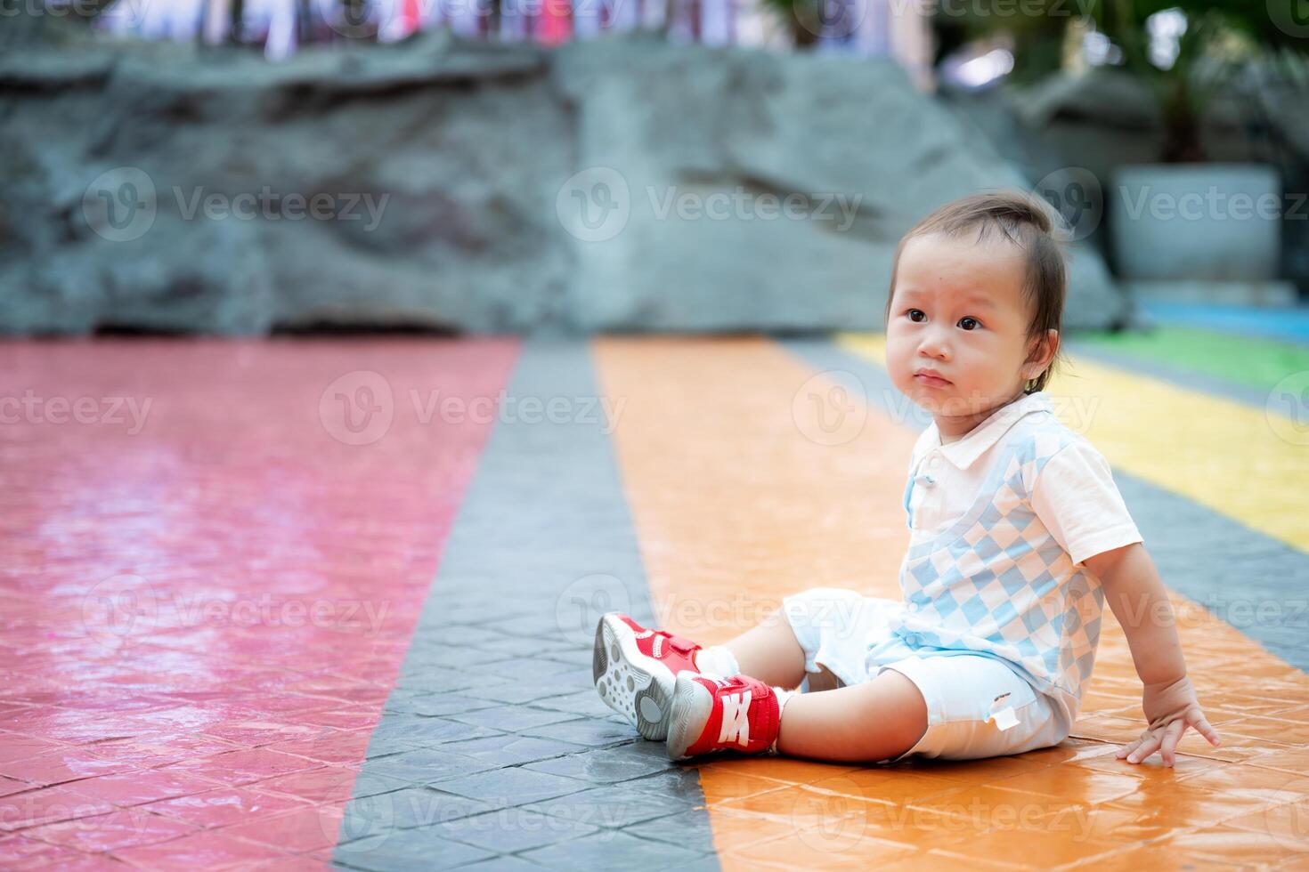 ritratto di carino asiatico bambino ragazzo è seduta su il pavimento a il terreno di gioco, contento bambino indossare blu e bianca Abiti e rosso scarpe da ginnastica, uno anno vecchio bambino. copia spazio. foto