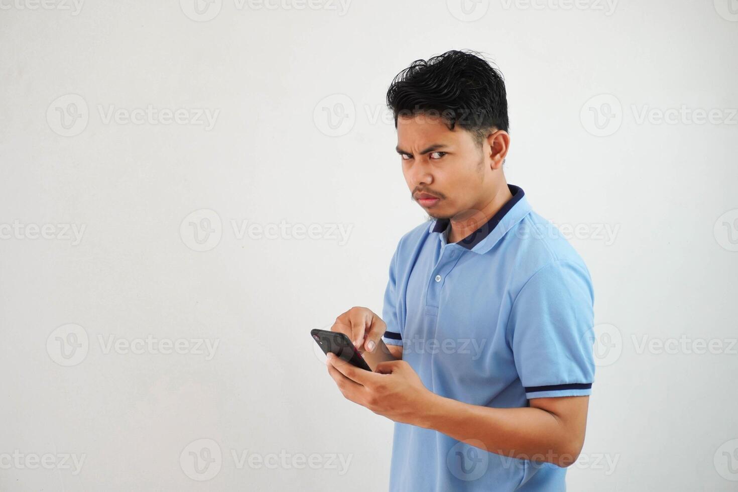 giovane asiatico uomo in piedi Tenere un' Telefono con un arrabbiato e disapprovazione espressione indossare blu t camicia isolato su bianca sfondo foto