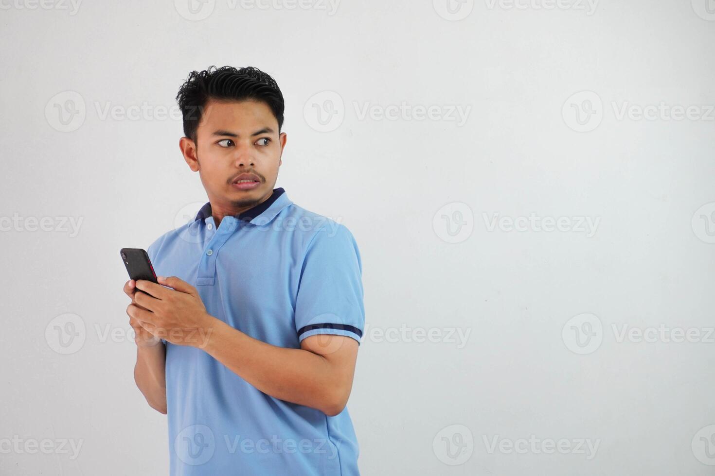 sorpreso in piedi giovane asiatico uomo indossare blu t camicia con Tenere smartphone, provando per schivare isolato su bianca sfondo foto