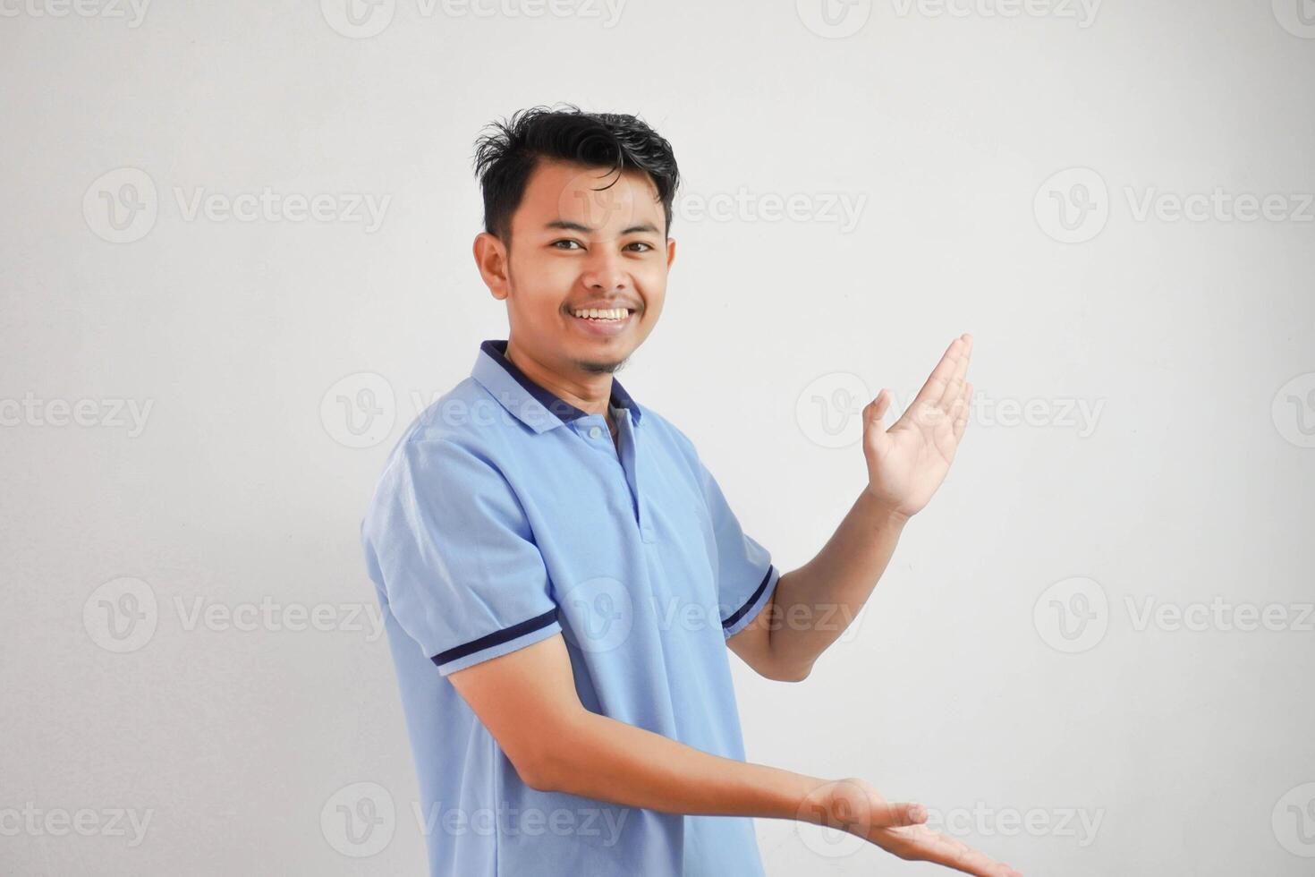 sorridente asiatico uomo Aperto braccia puntamento per il lati indossare blu t camicia isolato su bianca sfondo foto