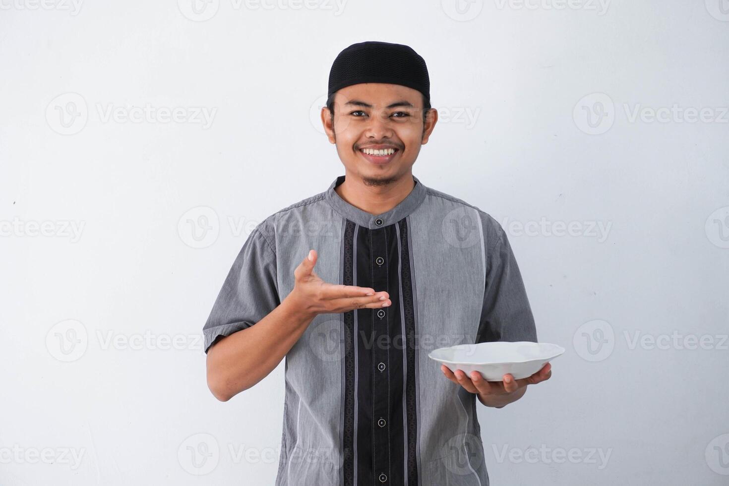 sorridente o contento giovane asiatico musulmano uomo mostrando eccitato espressione mentre Tenere vuoto cena piatto indossare grigio musulmano Abiti isolato su bianca sfondo foto