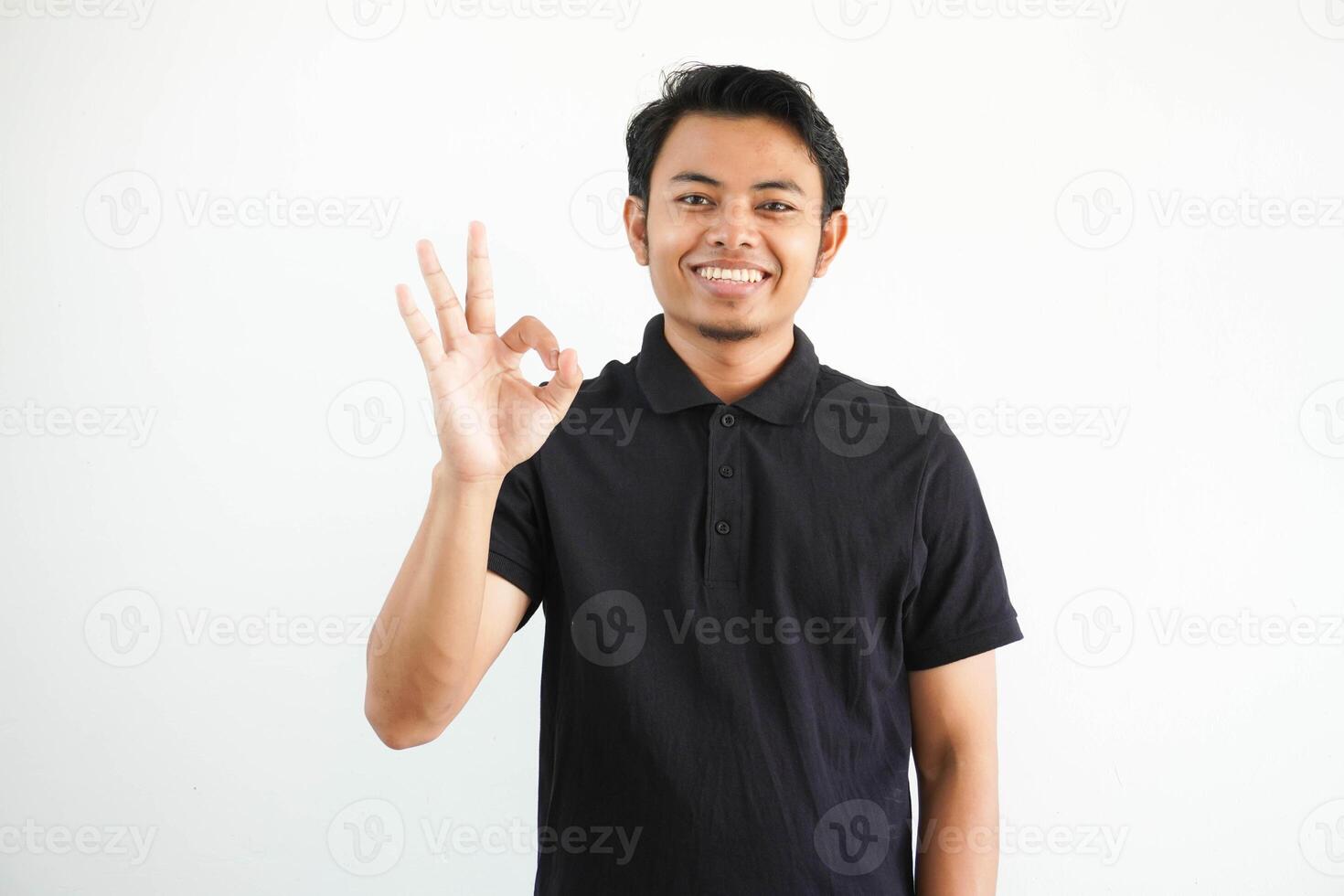 giovane asiatico uomo sorridente amichevole mentre dando ok dito cartello indossare nero polo t camicia isolato su bianca sfondo foto