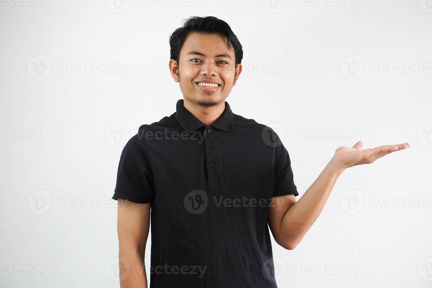 sorridente giovane asiatico uomo nel nero polo camicia, bianca fondale mostrando un' copia spazio su un' palma e Tenere un altro mano su vita. foto