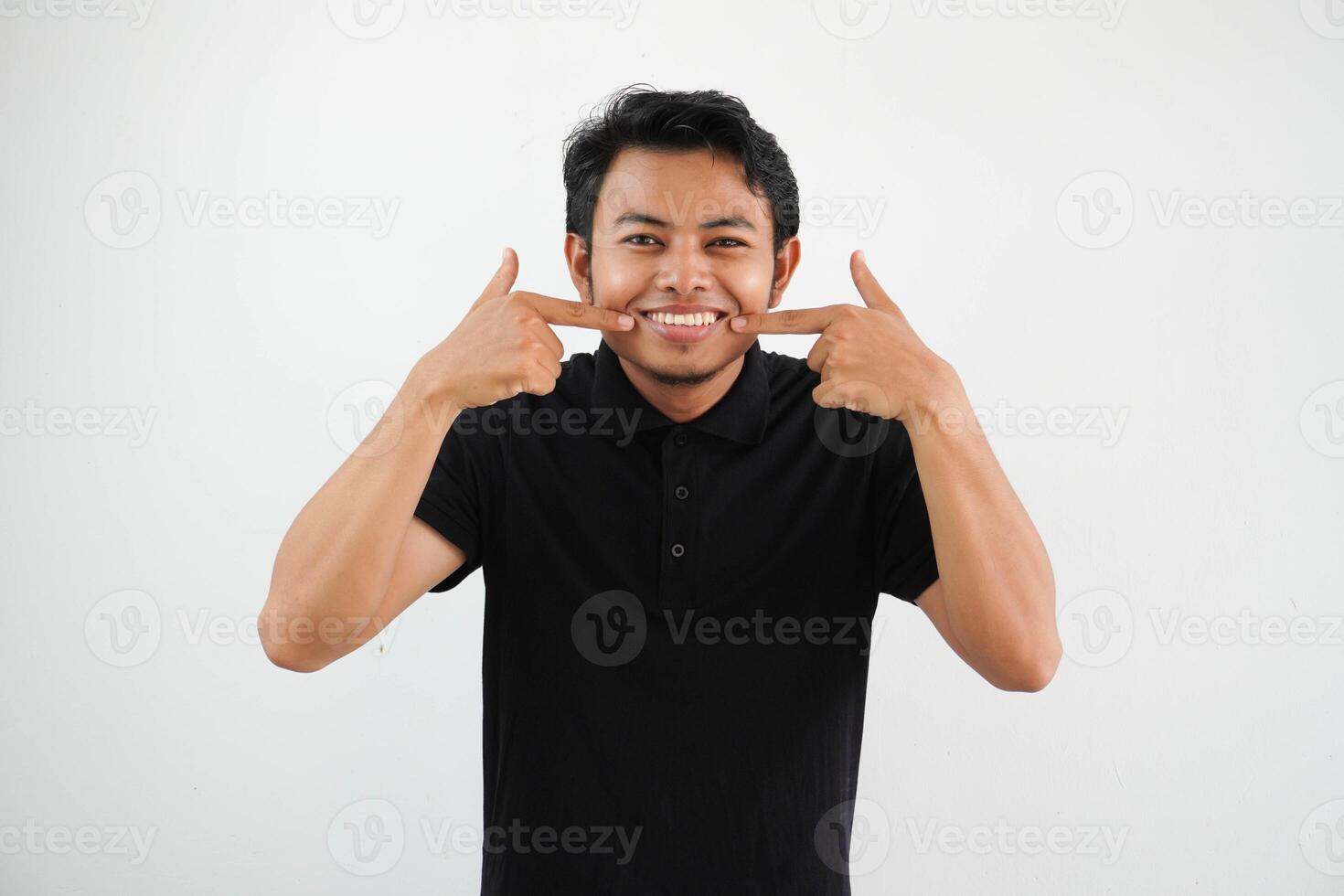 sorridente giovane asiatico uomo puntamento per il suo bocca con contento espressione indossare nero polo t camicia isolato su bianca sfondo foto