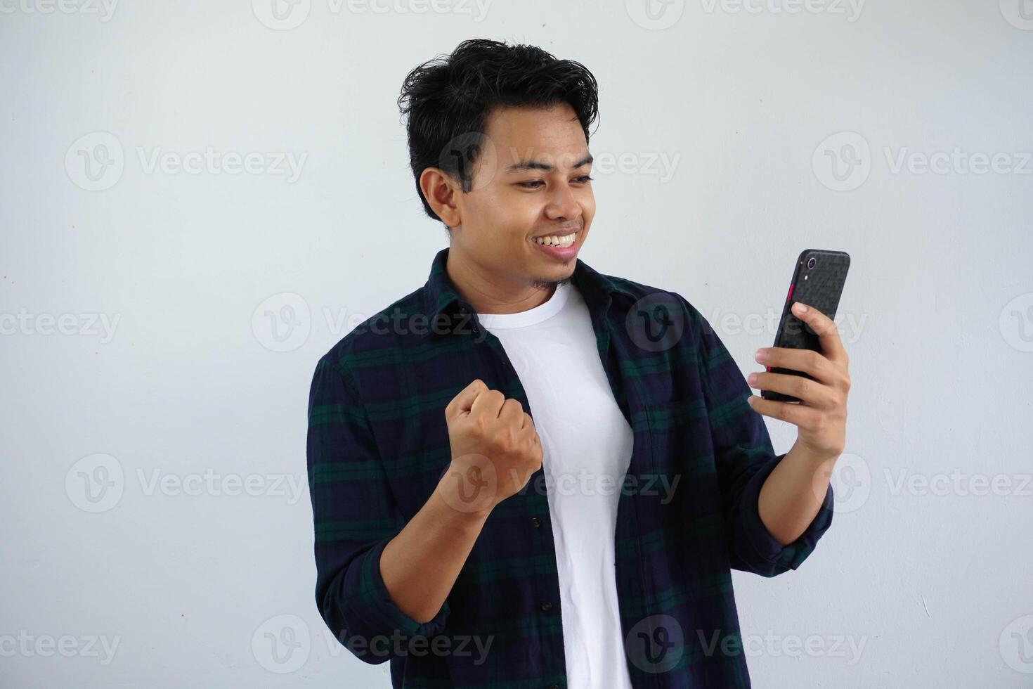 giovane asiatico uomo stringendo cazzotto mostrando eccitazione quando guardare per il suo Telefono isolato su bianca sfondo foto