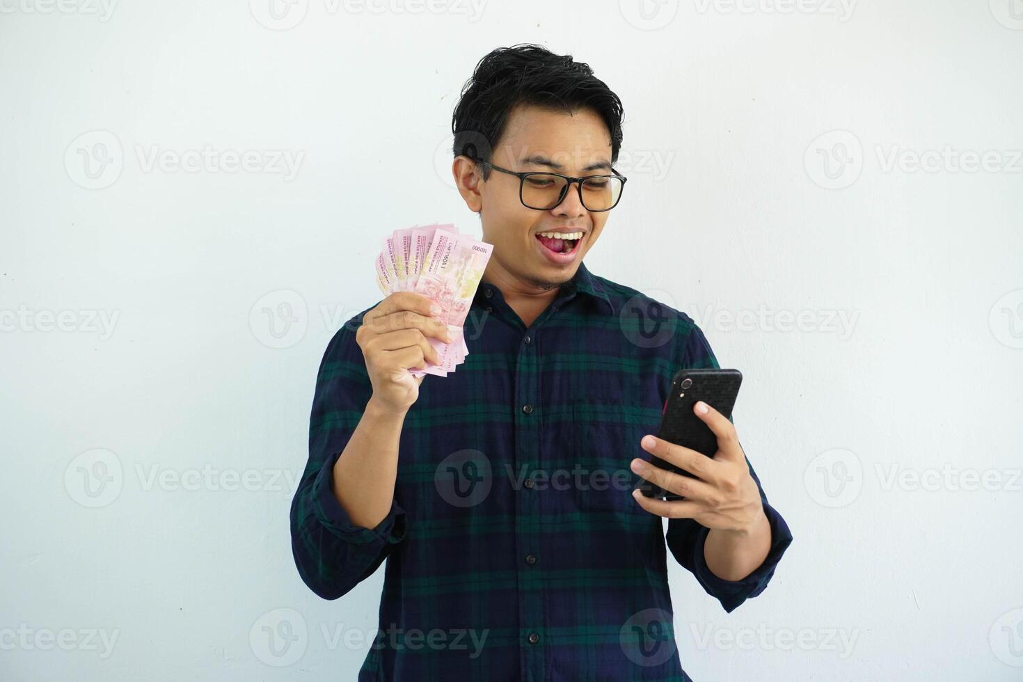 giovane asiatico uomo mostrando sorpreso espressione quando guardare per il suo mobile Telefono mentre Tenere carta i soldi isolato su bianca sfondo. foto