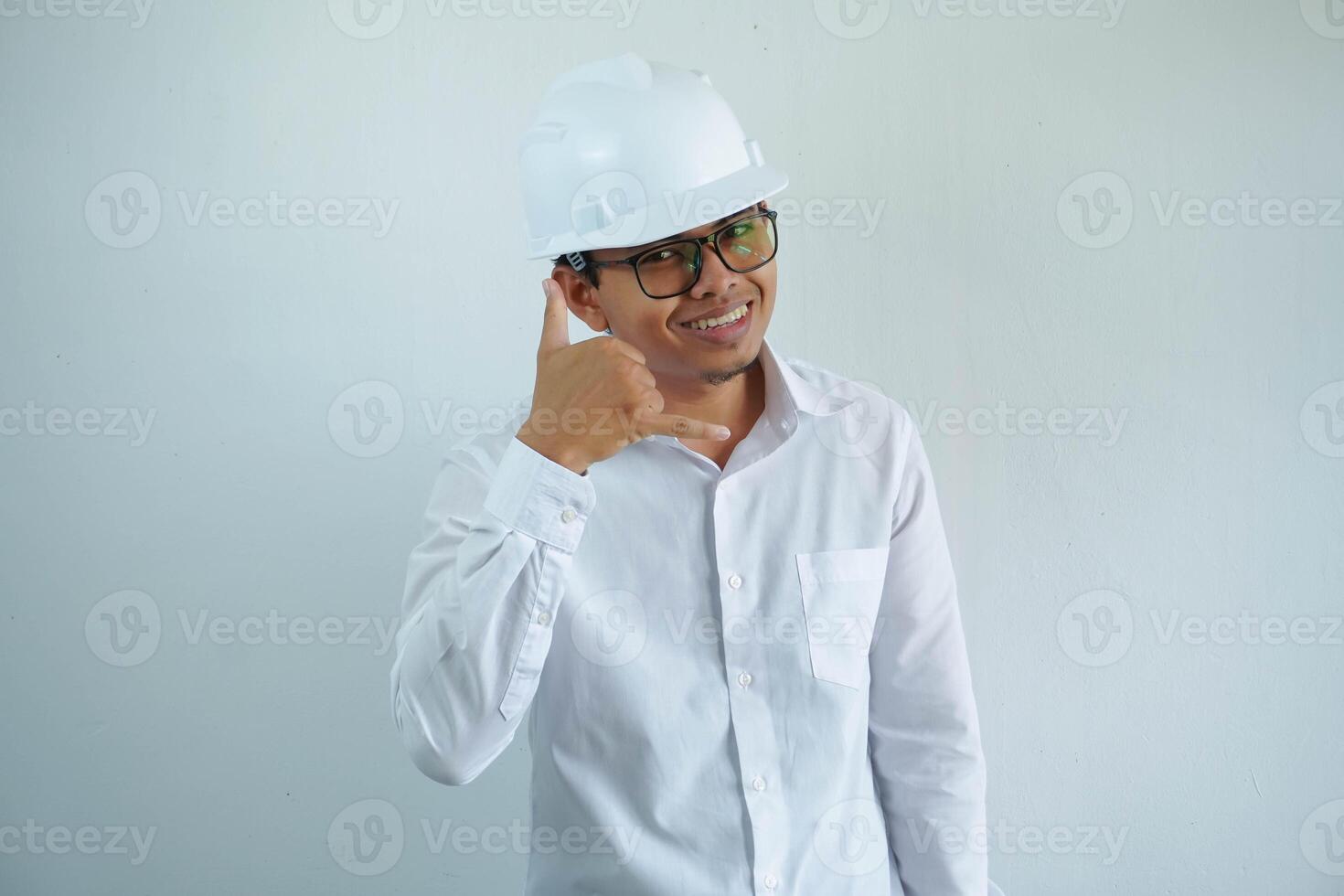 giovane asiatico uomo è ingegnere indossare casco in piedi con mostrando un' mobile Telefono chiamata gesto con dita e sorridente con fiducioso, architetto o contraente, lavoratore o lavoro duro e faticoso, industriale concetto. foto