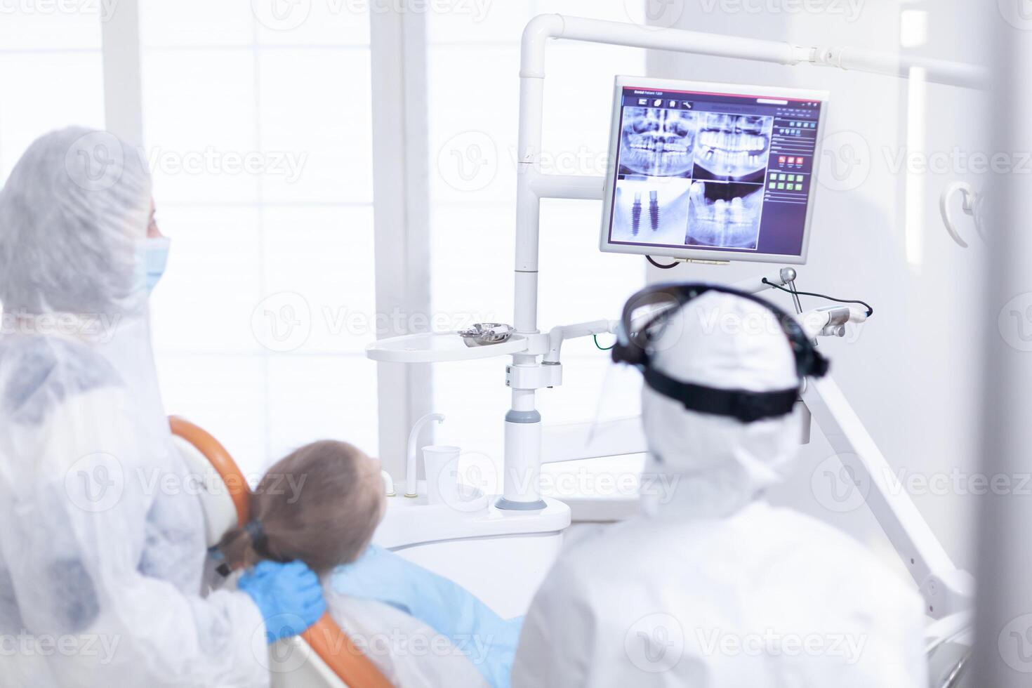 ortodontista analizzando digitale radiografia durante ragazzo paziente visitare vestito nel pp. stomatologia nel protezione completo da uomo per coroanvirus come sicurezza precauzione guardare a digitale bambino denti raggi X durante consultazione. foto