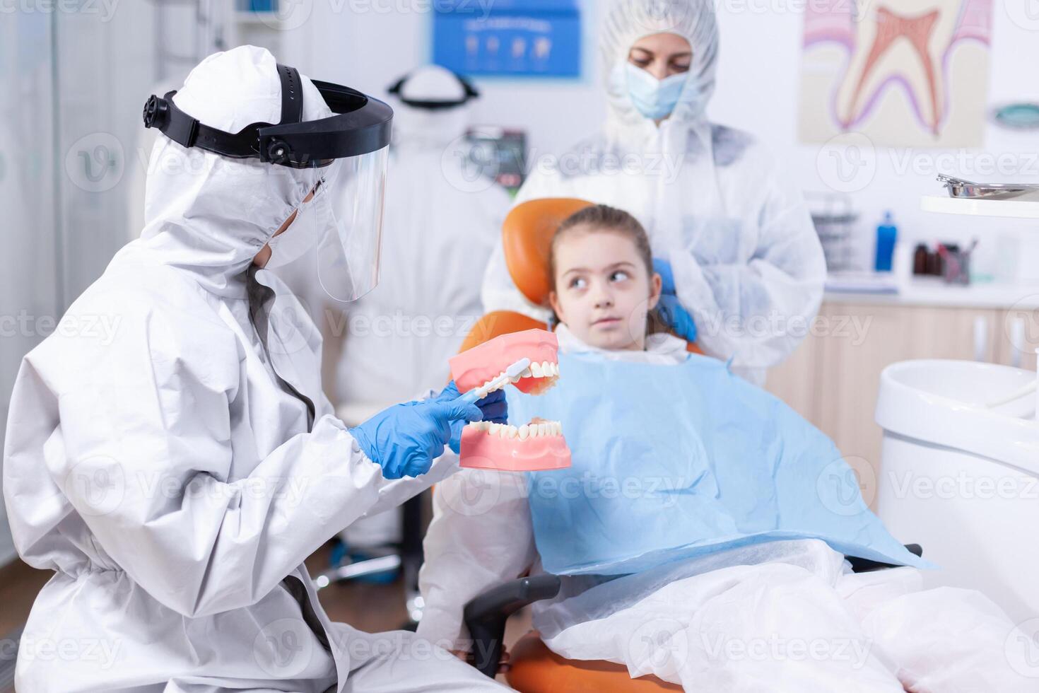 dentista insegnamento ragazzo Come per spazzola denti vestito nel pp. poco ragazza nel ppe completo da uomo durante covid19 e madre ascoltando stomatologia parlando di dente hygine nel odontoiatria clinica Tenere mascella modello. foto