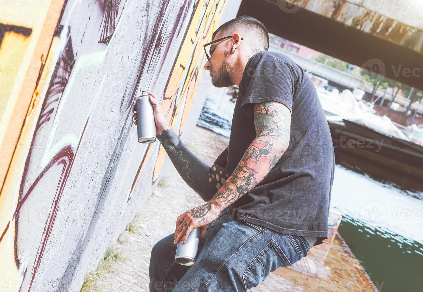 graffiti artista pittura con un' colore spray può su il parete nel il città - giovane tipo fare un' murale Il prossimo per il fiume - urbano e strada arte stile di vita concetto foto