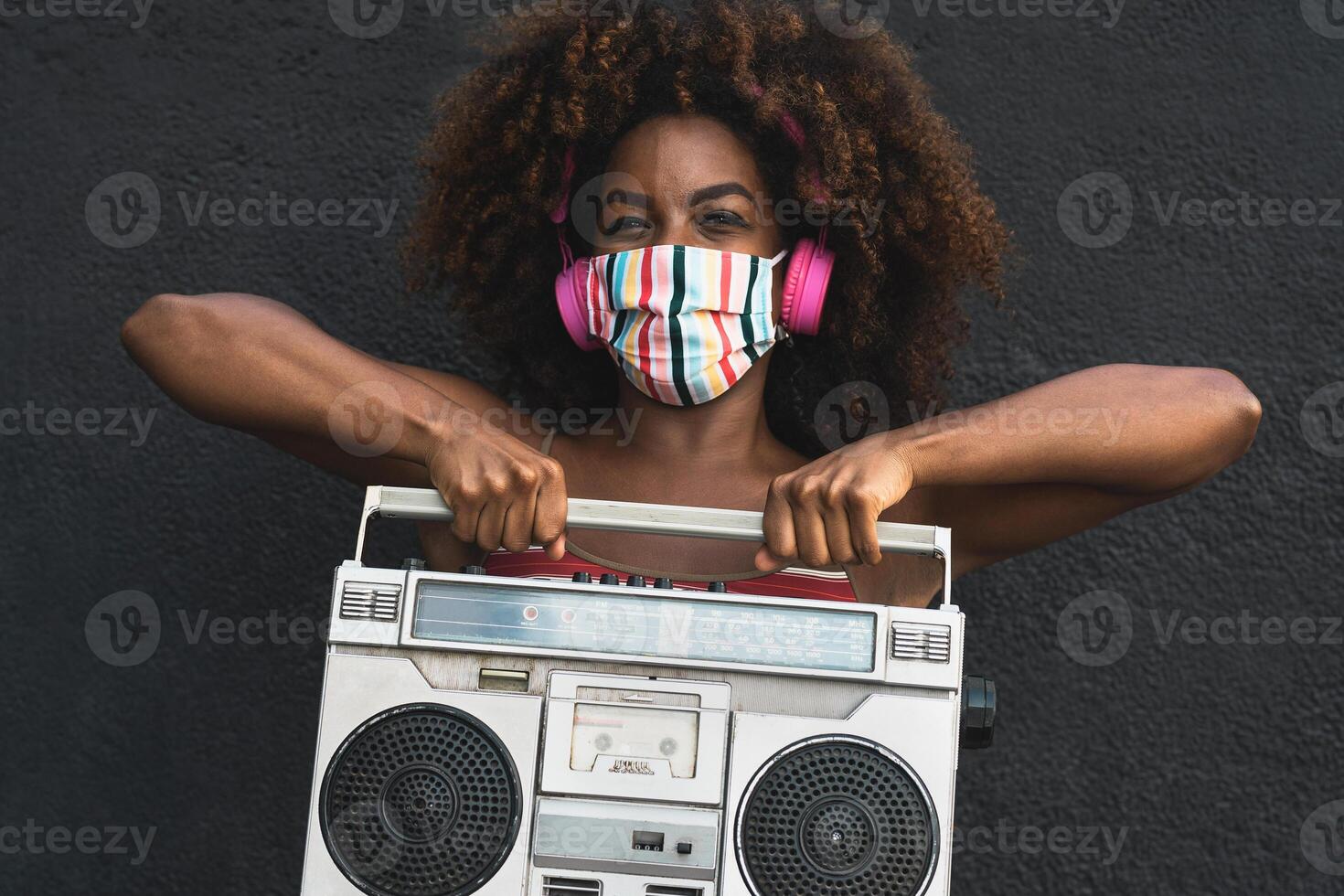 giovane afro donna indossare viso maschera danza all'aperto mentre ascoltando per musica con senza fili cuffie e Vintage ▾ Boombox stereo foto
