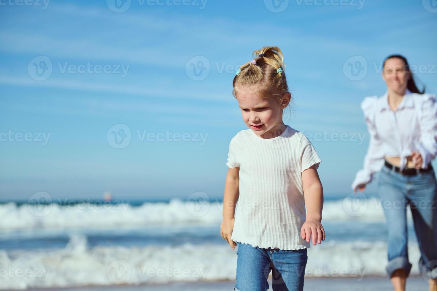 pensieroso bionda bambino ragazza 2-3 anni vecchio nel bianca maglietta e casuale denim, a piedi su il spiaggia con sua contento madre foto
